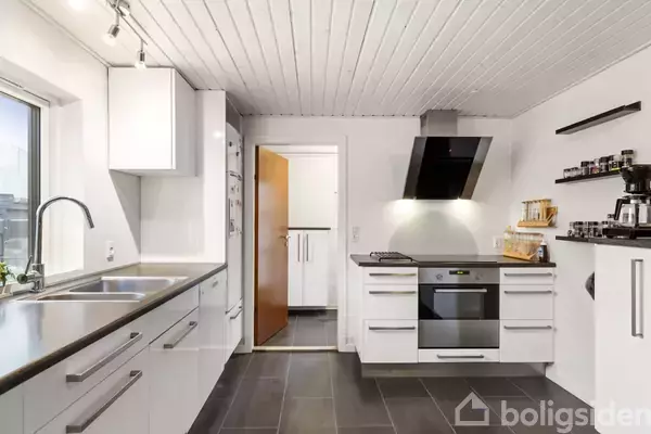 Hvidt køkken med sorte fliser, hvor en ovn og kogeplade er monteret under en emhætte. Til højre står hylder med køkkenapparater. Dør fører til tilstødende rum.