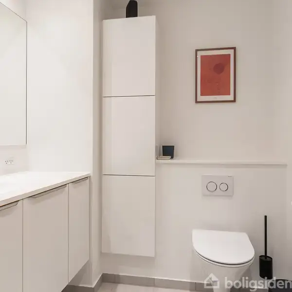 Badeværelse med hvidt interiør, vask til venstre, væghængt toilet til højre, og en høj skabsenhed. En enkelt billedramme hænger over toilettet, og der er flisegulv.