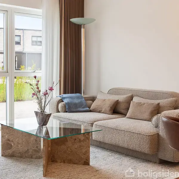 En beige sofa står i en lys stue ved siden af et glasbord med blomsterdekoration. Store vinduer lader naturligt lys ind, og udenfor ses en modernistisk bygning og grønne planter.