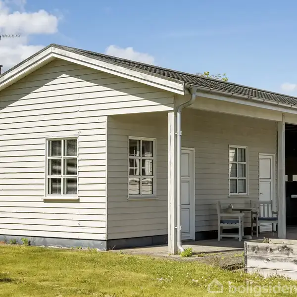Hvidmalet træhus står stille med en veranda, to hvide døre og vinduer; en bænk placeret foran. Gule plæner omgiver huset, og himlen er klar blå med få skyer.