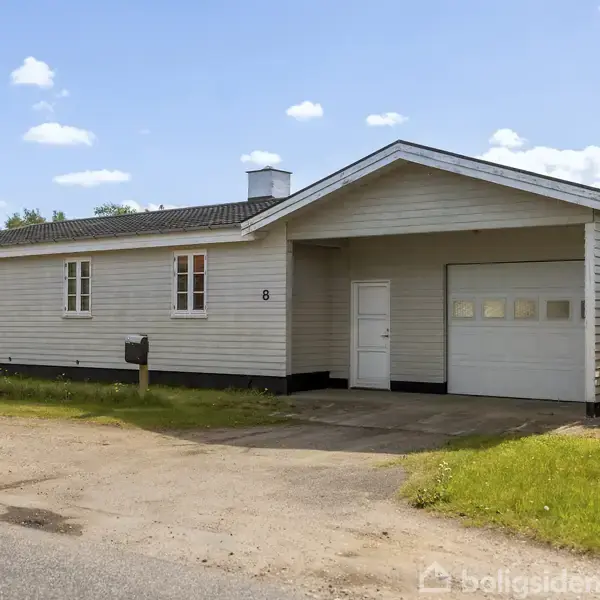 Et hvidt enfamiliehus med en fladt tag og tilhørende garage står ved en grusvej i et roligt kvarter. Adressen "8" ses ved døren. Græsplæne omgiver huset.