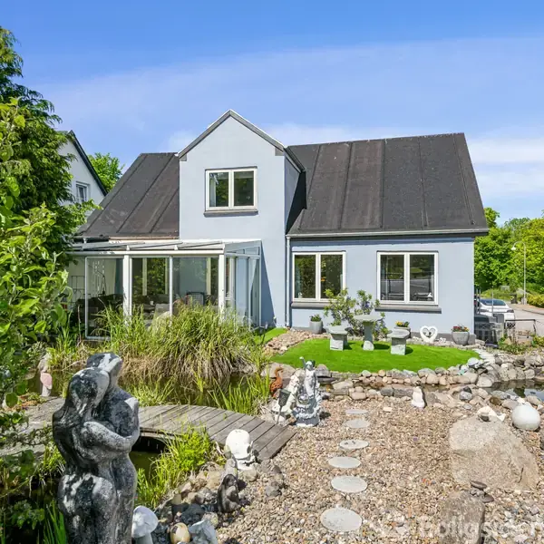 Et gråt hus med sorte tage står i en velplejet have med stier, en lille dam og stenskulpturer, omkranset af frodig vegetation og blå himmel.
