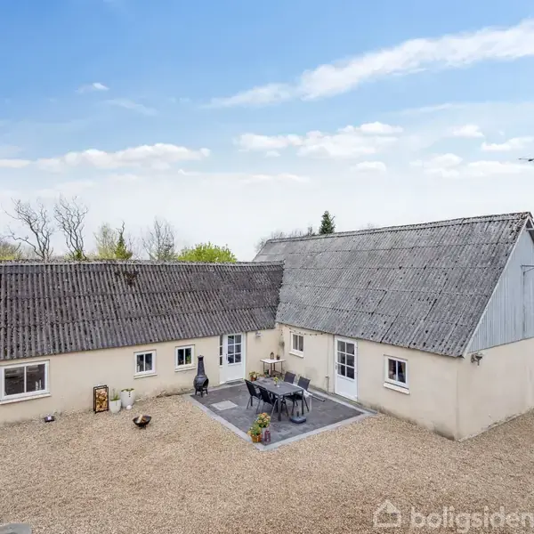 Et hvidt hus med skråt tag står i en grusbelagt gårdsplads. Der er en terrasse med sorte havemøbler, omgivet af træer og buske under en blå himmel med lette skyer.