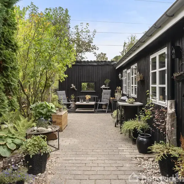 En brolagt gangsti fører gennem en frodig have med mange planter. Til højre ses et sortmalet hus med vinduer. I baggrunden er der et opholdsområde med borde og stole.