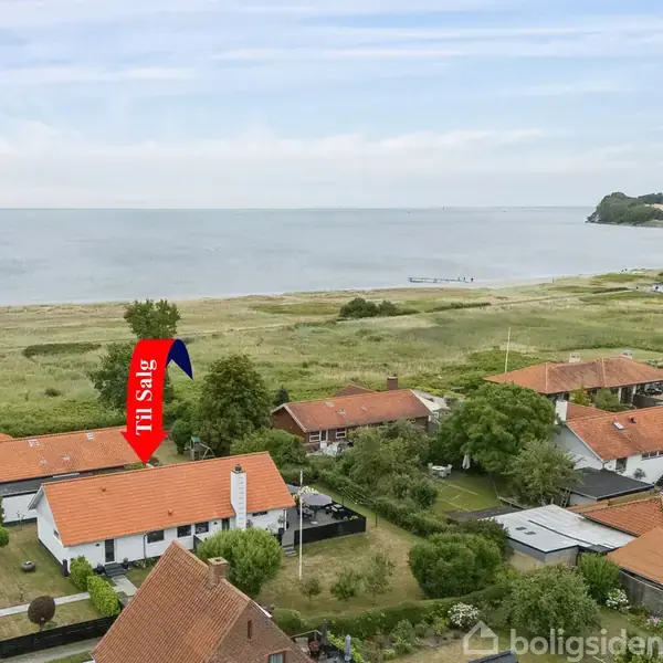 Hus til salg, markeret med rød pil, omgivet af andre huse med røde tage tæt ved stranden. Strand og vand udgør baggrunden, mens grønne områder omkredser husene. Tekst: "Til Salg".