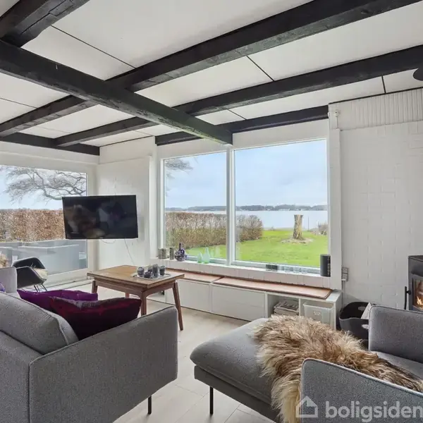 En moderne stue med grå sofaer og brændeovn. Store vinduer giver udsigt til en grøn have og sø. Fjernsyn og sofabord er placeret mellem møblerne.