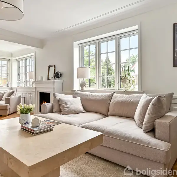 En beige sofa står foran et stort vindue, omgivet af hvide vægge og træmøbler. Et sofabord er fyldt med bøger og planter. Stuen har et lyst, moderne design.