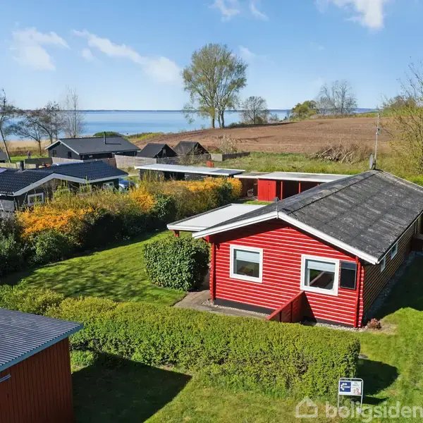 En rød træhytte med sort tag står omgivet af en grøn have og tætklippede hække. Flere huse ses i baggrunden med udsigt til et kystlandskab og havet.