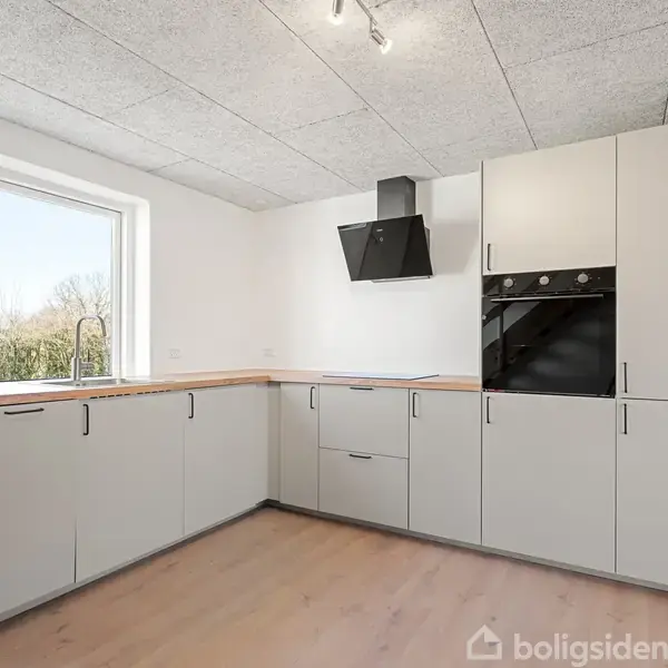 Et moderne køkken med hvide skabslåger og en sort indbygget ovn. Der er et stort vindue, der giver udsigt til en have med træer. En loftslampe lyser rummet op.