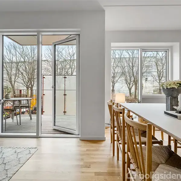 Spisebord med træstole står i et moderne spisekøkken med lyse trægulve. Glasdøre fører ud til en balkon med træmøbler og udsigt til træer udenfor.