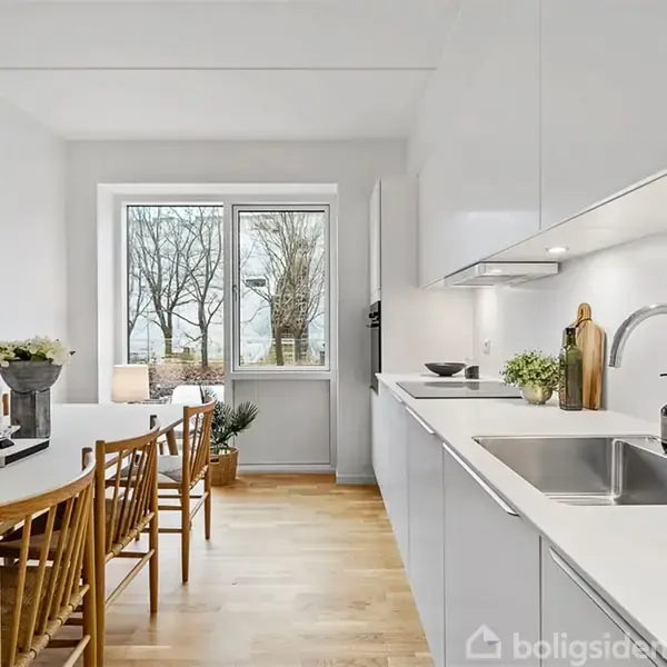 Et moderne køkken med hvide skabe og skinnende bordplade. Et langbord med træstole står ved vinduet, som åbner ud til træer udenfor, skaber en lys og indbydende atmosfære.