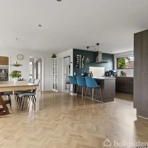 Køkken-alrum med et moderne træbord og stole i forgrunden. Tre blå barstole står ved en mørk køkkenø. Hvidvægget rum med parketgulv og udgang til andre rum på venstre side.