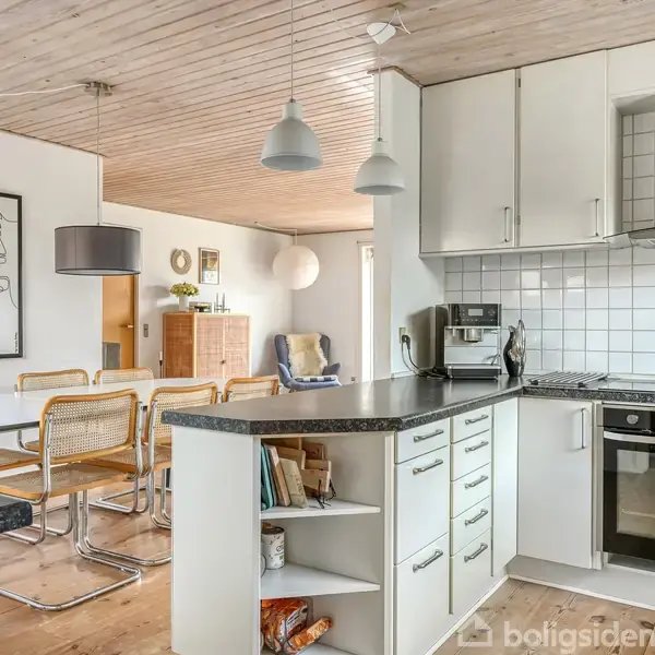 Køkken med hvide skabe og sort bordplade, integreret ovn og kogeplade. Åben til spiseområde med bord, stole og vægkunst. Trægulve og lofter skaber en varm atmosfære.