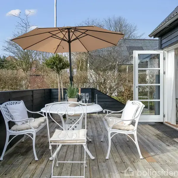En terrasse med hvide havemøbler står under en beige parasol. Døren til huset i baggrunden er åben, og grønne buske omkranser området.