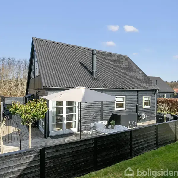 Et sortmalet hus med skråt tag har en tilhørende terrasse, hvor der står et parasol med havemøbler. Huset er omgivet af en hæk og en velplejet græsplæne.