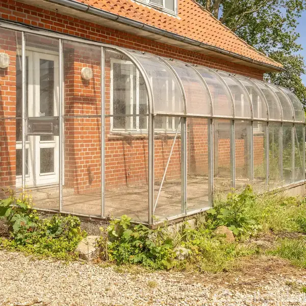 Væksthus tilknyttet rød murstensvilla, har buede glasrammer indrammet af metal. Omgivet af grusbelagt område og grønne planter.