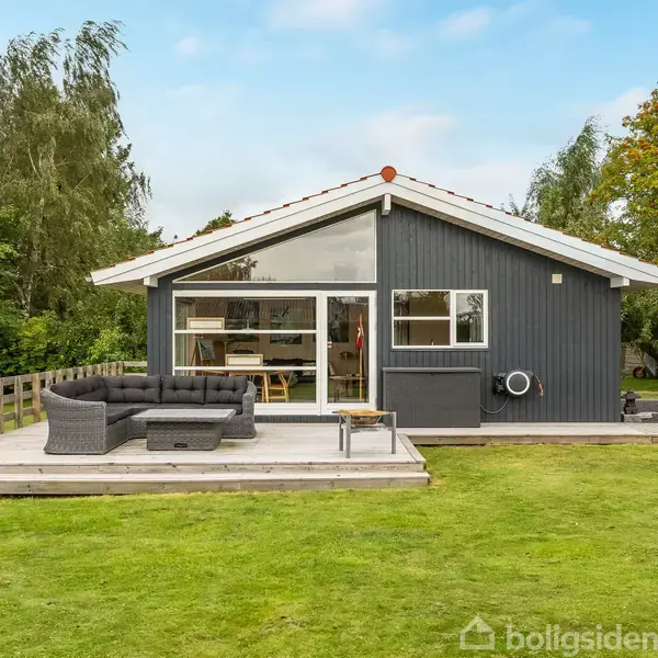 Et lille, moderne sommerhus med sort træbeklædning står på en græsplæne. Der er en terrasse med udendørs sofa og træbord. Træer omgiver huset.