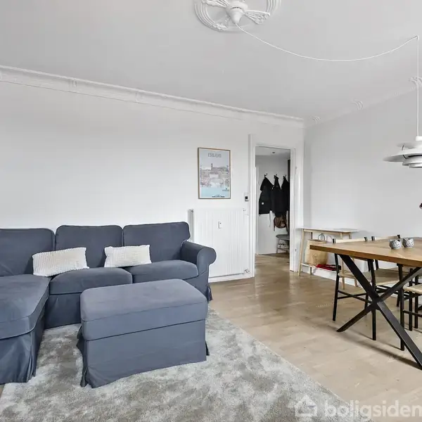 En mørkeblå sofa står i en lys stue med trægulv, ved siden af et spisebord med sorte stole. En stående lampe og en vase med blomster er placeret i rummet.