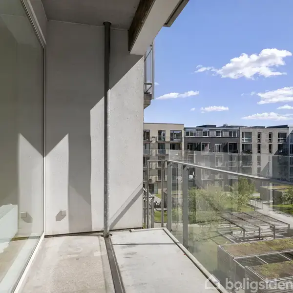 En balkon med glasrækværk tilbyder udsigt over en moderne boligkompleks. Skinnende vinduer reflekterer sollys, mens skyer svæver på en blå himmel, hvilket skaber en lys atmosfære.