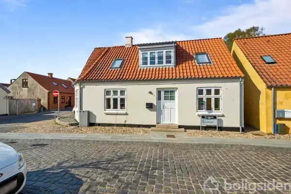 Et lille hus med rødt tegltag står langs en brostensbelagt vej i et bymiljø. Et skilt foran huset viser teksten: "Til salg" og "Estate".