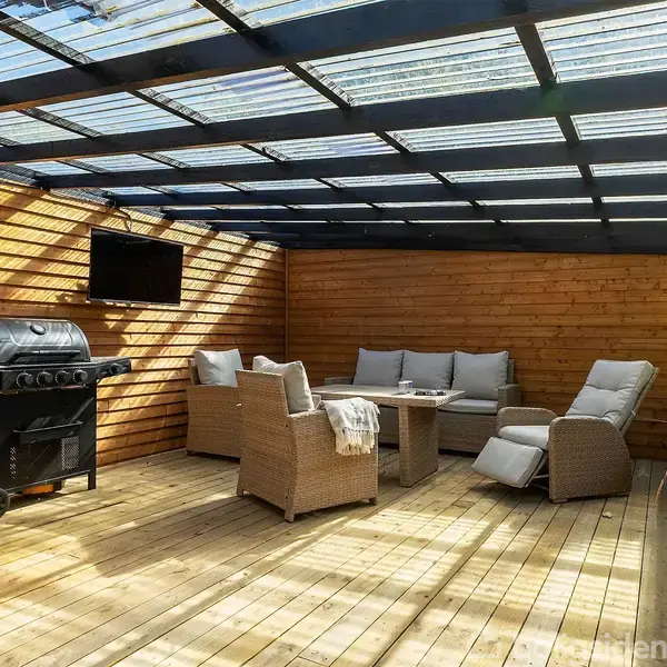 Grill står på en overdækket træterrasse ved siden af kurvestole og et sofabord. En væghængt tv-skærm hænger ved siden af grillen. Terrasse er omkranset af trævægge.
