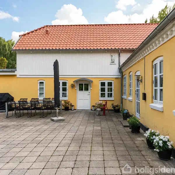 Huset er gult med hvide vinduer og har en rød tag. Foran er en terrasse med havemøbler og grill. Plankerne er lysegrå, og blomster i potter står langs væggen.