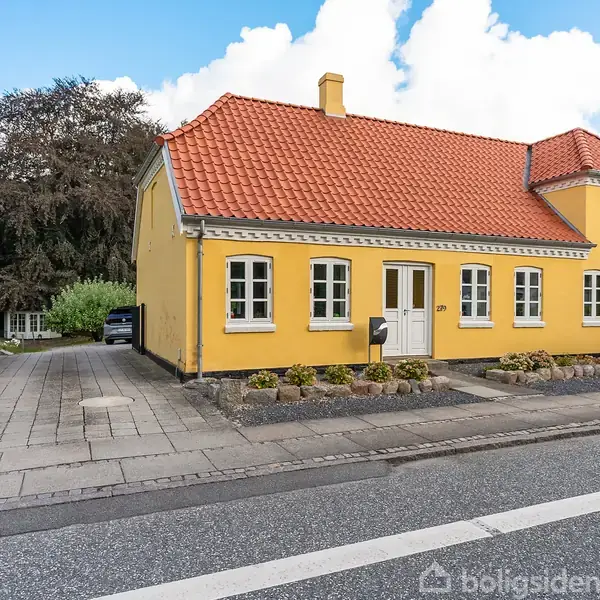 Et gult hus med rødt tegltag står langs en vej. Indkørslen er til venstre omgivet af grønne buske og træer. Huset har flere hvide vinduer og dør.