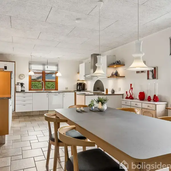 Køkkenbord er omgivet af træstole, hvor en vase med blomster står centralt. Omkring er hvide skabe, vinduer med udsigt til en grøn have, og moderne lamper hænger fra loftet.
