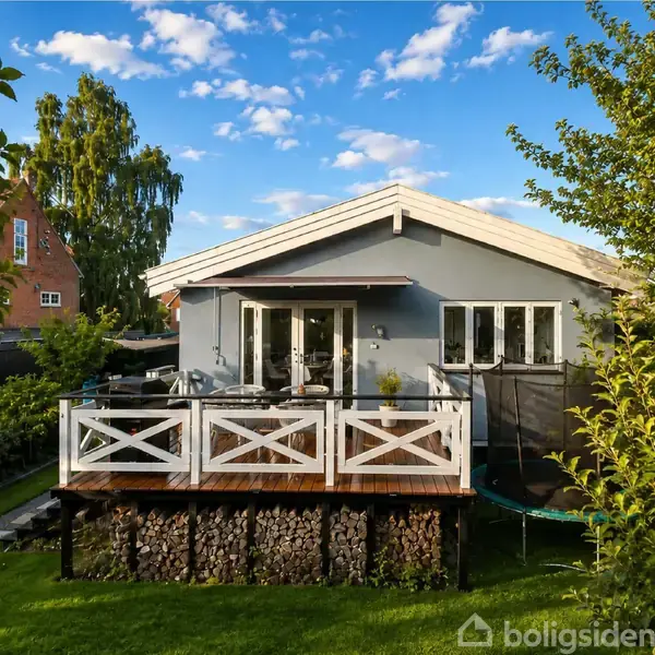 Blåt hus med terrasse, hvor der står havemøbler. Foran er der en velplejet have med træer og buske, under en blå himmel med lette skyer.