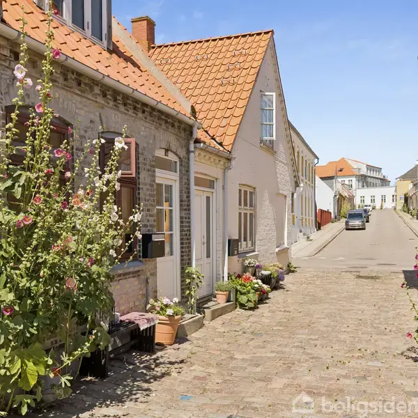Gamle huse med orange tegltage står langs en brolagt gade. Blomster og planter pryder facaderne. I baggrunden ses flere bygninger under en klar blå himmel. Ingen tekst er til stede.