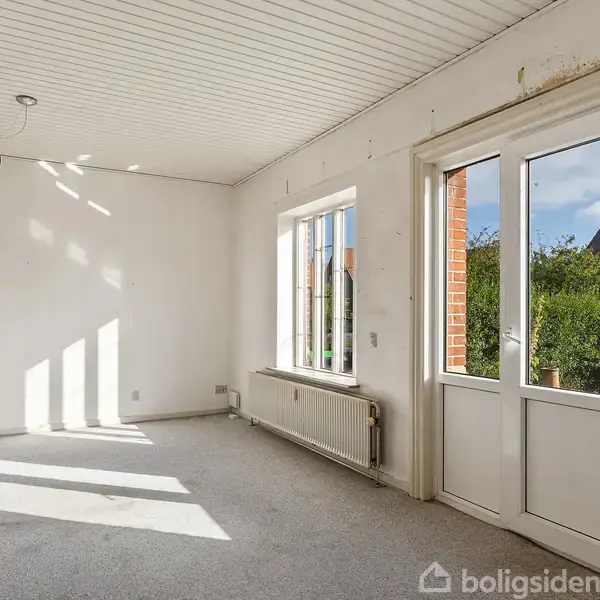 Køkken med lyse skabe og træbordplade. Ovn og vask integreret. En tæppe ligger på gulvet. To planter og sæbedispenser står på bordet. Store vinduer til højre.