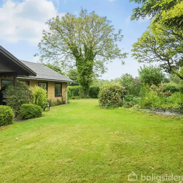 Hus med brun mursten står stille i en velholdt have med frodigt græs og forskellige buske og træer omkring. I baggrunden ses en stor, grøn trækrone mod en blå himmel.