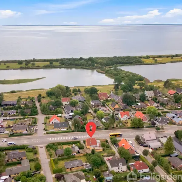 Byhus markeret med en rød pin fra oven, omringet af flere huse og grønne områder. I baggrunden ses en vej og en kystlinje med vand, under en blå himmel.