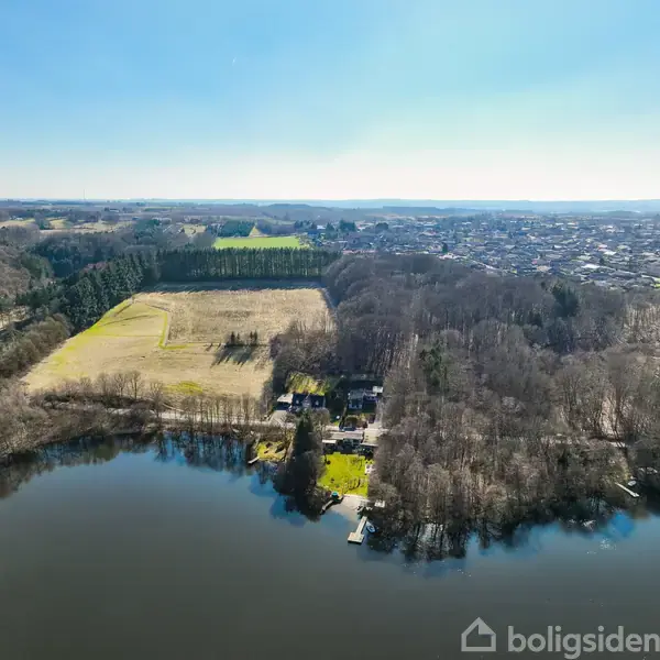 Sø med spejlblank overflade ligger i forgrunden. Huse er omgivet af skov og åbne marker. I baggrunden ses en by med spredte bygninger under en klar himmel.