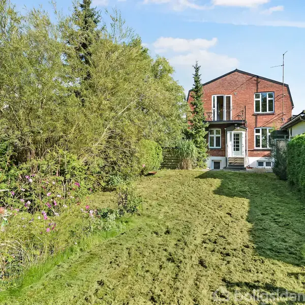 Et murstenshus med en blå dør og hvide vinduer står i baggrunden. En nyslået græsplæne omgivet af buske og træer fylder forgrunden. Ingen tekst til stede.