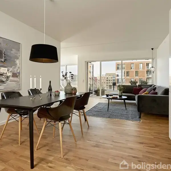 Spisebord med sorte stole stående i en moderne stue med trægulv. En sort lampe hænger over bordet. Store vinduer slipper lys ind, og i baggrunden ses et opholdsområde med sofa.