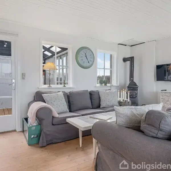 Stue med grå sofa, sofabord og væghængt fladskærm. Brændeovn i hjørnet. Vægure over vinduet. Lys fra verandadør og vinduer. Lys og indbydende atmosfære.