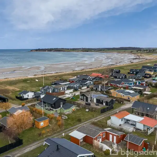 Kysthuse ligger spredt langs en sandstrand ved en rolig kystlinje. Husene står på grønne grunde, mens havet strækker sig ud mod horisonten under en delvist overskyet himmel.
