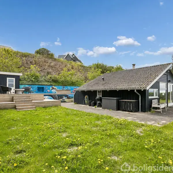 Sommerhus med sort træfacade og skråt tag står på en grøn græsplæne. Til venstre er en forhøjet træterrasse. Baghaven er omgivet af træer og buske under en blå himmel.
