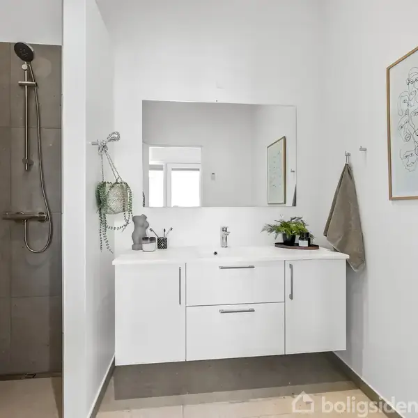 Håndvask med hvidt skab og spejl på væggen, flankeret af en bruser i bruseniche til venstre; billedet hænger til højre på en hvid badeværelsesvæg.