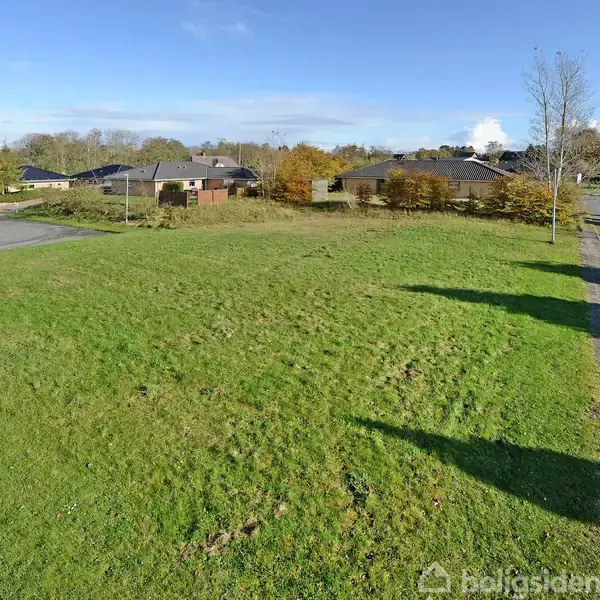 Grønt græsareal ligger stille ved siden af en asfalteret vej i et boligkvarter med huse i baggrunden. Himlen er blå, og solen skinner lyst.