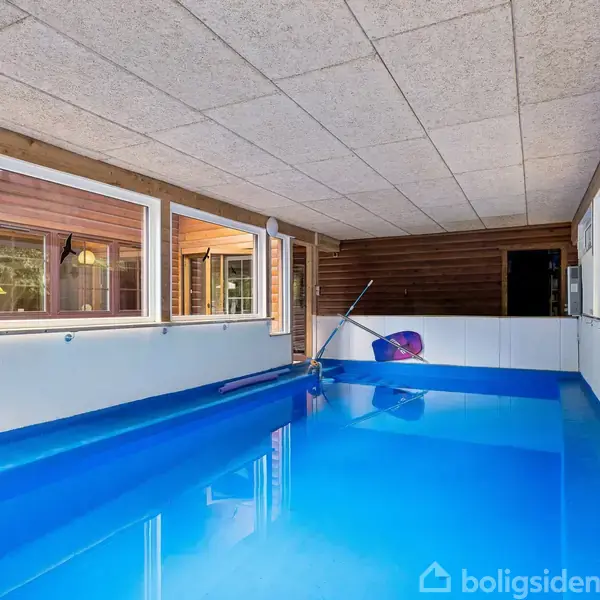Indendørs swimmingpool med klart blåt vand og en flydende badedyrsring. Poolen er omgivet af store vinduer, der giver udsigt til træbeklædte vægge og en grøn have udenfor.