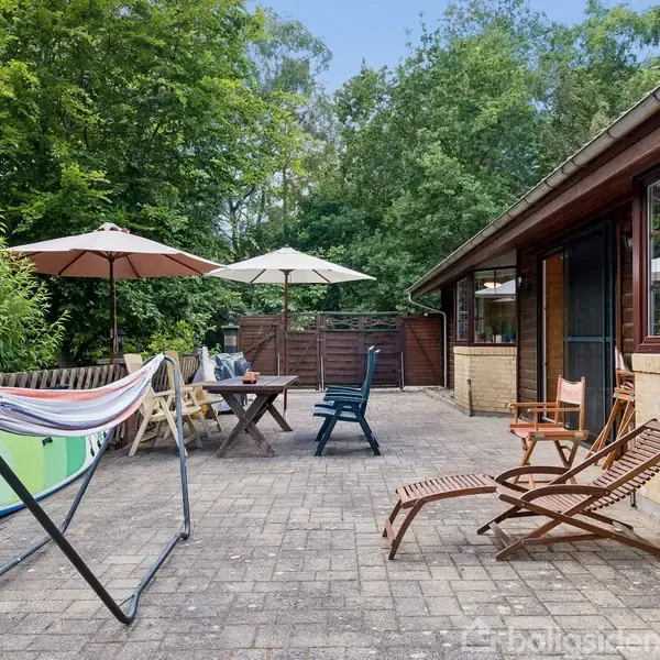 Hængekøje hænger på et stativ på en flisebelagt terrasse foran et hus. Ved siden af står liggestole, parasoller og et bord. Træer omkranser området.