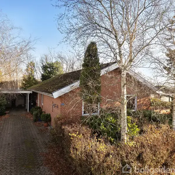 Et murstenshus med skrånende tag omkranset af træer og buske. Indkørslen er brolagt og fører til en carport ved siden af huset.