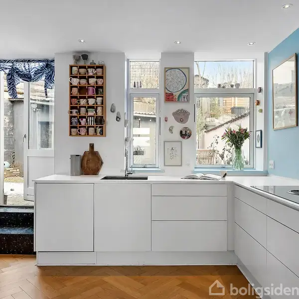 Køkkenbord med hvide skuffer står i et rummeligt køkken. Billeder og dekorationer hænger på en lyseblå væg. Store vinduer slipper lys ind. Trappe fører til en udendørs dør.