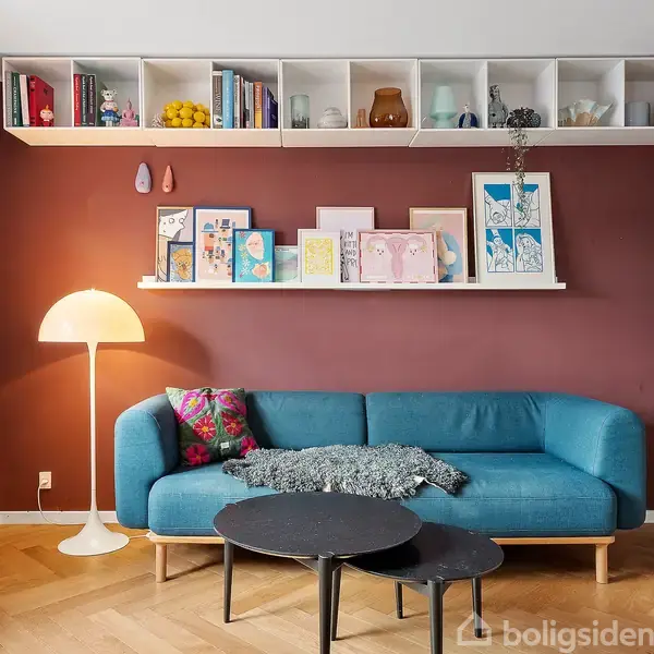 En blå sofa står mod en mørkerød væg med kunstværker og en bogreol. En stålampe lyser ved siden af sofaen. Trægulv og et farverigt billede hænger på væggen.