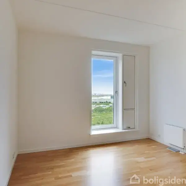 Et tomt rum med trægulv, hvor et åbent vindue viser udsigt til det fjerne landskab. Væggene er hvide, og der er en radiator under vinduet.
