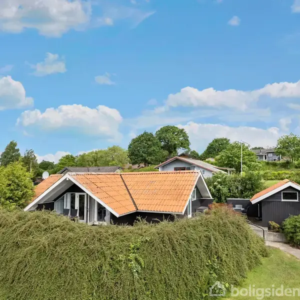 Rødt tegltagshus omgivet af tæt grønt hegn, beliggende i frodigt landskab med andre huse i baggrunden under en delvist skyet blå himmel.
