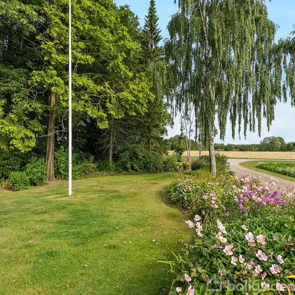 En græsplæne med blomsterbede fyldt med farverige blomster i forgrunden. En flagstang står blandt store træer. En grusvej fører til marker, med en lille rød bygning i baggrunden.