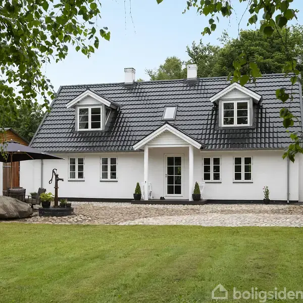 Hus står stille med sorte tagsten, hvide vægge og to kviste. Foran huset er en græsplæne, indkørsel af brosten, og træer omgiver bygningen.